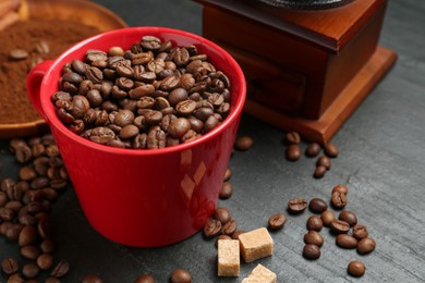 Photo of Aromatic coffee beans in cup, grinder and brown sugar on black table, closeup
