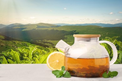 Image of Green tea in glass teapot, mint leaves and lemon on table against tea plantation in mountains. Space for text