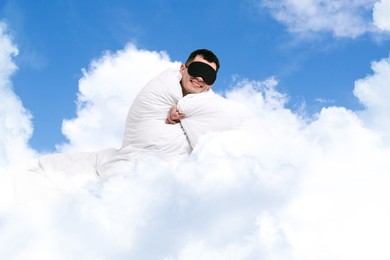 Image of Happy man with blanket and sleep mask sitting on fluffy cloud in blue sky. Bedtime