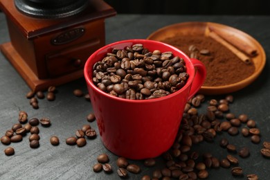Photo of Aromatic coffee beans in cup and grinder on black table, closeup