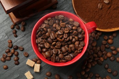 Photo of Aromatic coffee beans in cup, grinder and brown sugar on black table, flat lay