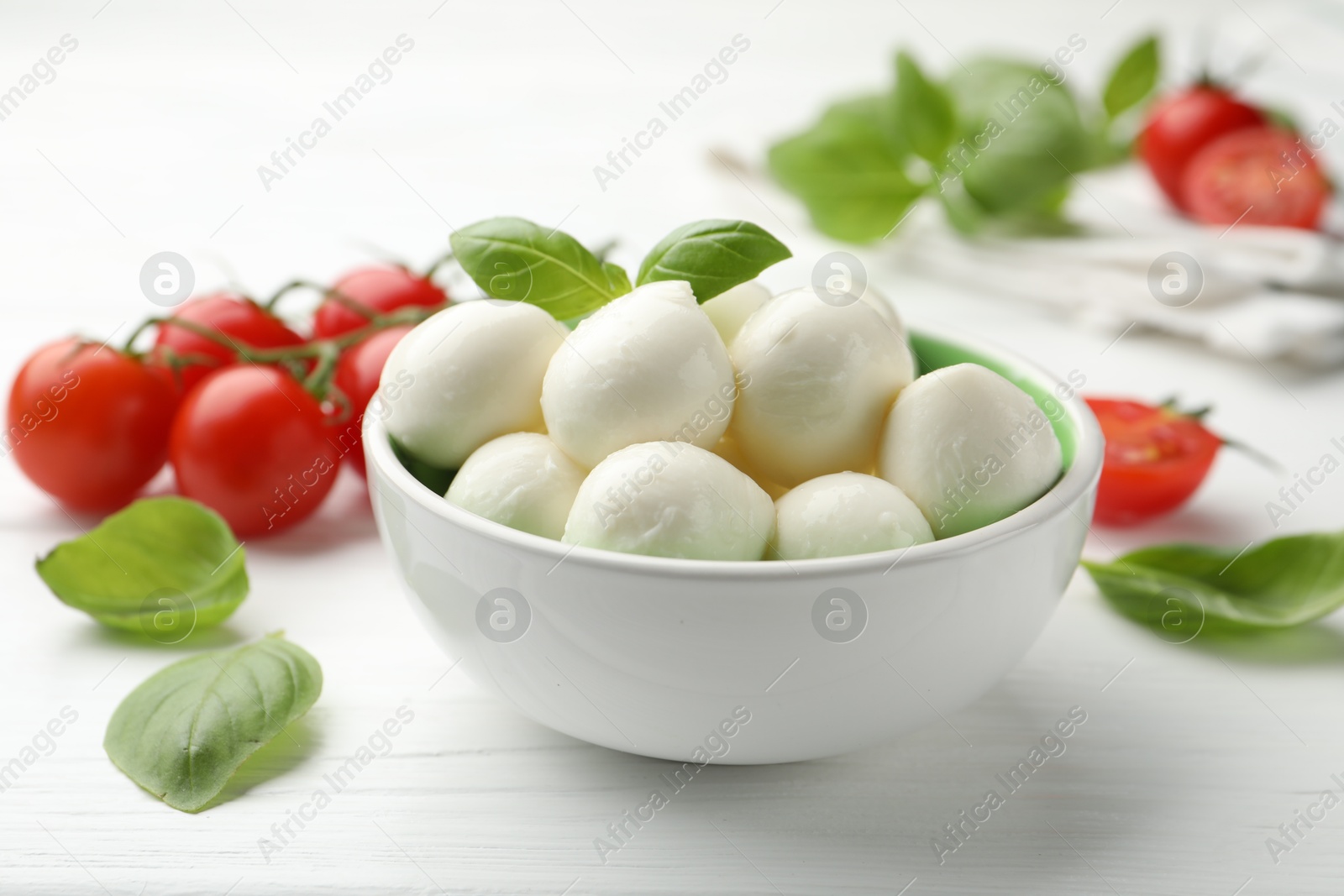 Tasty mozzarella cheese balls in bowl, basil and tomatoes on white wooden table, closeup Photo of Tasty mozzarella cheese balls in bowl, basil and tomatoes on white wooden table, closeup