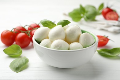 Photo of Tasty mozzarella cheese balls in bowl, basil and tomatoes on white wooden table, closeup