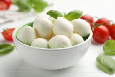 Photo of Tasty mozzarella cheese balls in bowl, basil and tomatoes on white wooden table, closeup