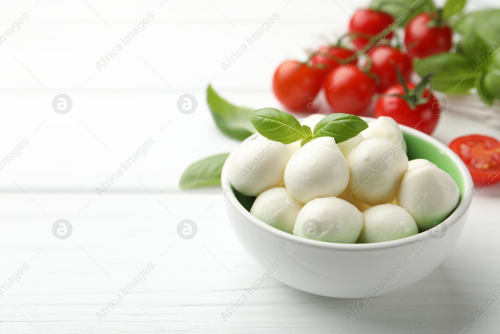 Tasty mozzarella cheese balls in bowl, basil and tomatoes on white wooden table, closeup. Space for text Photo of Tasty mozzarella cheese balls in bowl, basil and tomatoes on white wooden table, closeup. Space for text