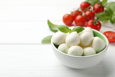 Photo of Tasty mozzarella cheese balls in bowl, basil and tomatoes on white wooden table, closeup. Space for text