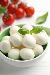 Photo of Tasty mozzarella cheese balls in bowl, basil and tomatoes on white table, closeup