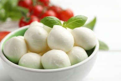 Photo of Tasty mozzarella cheese balls in bowl, basil and tomatoes on white table, closeup