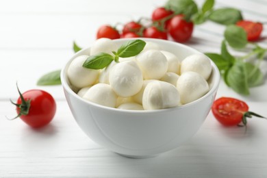 Photo of Tasty mozzarella cheese balls in bowl, basil and tomatoes on white wooden table, closeup