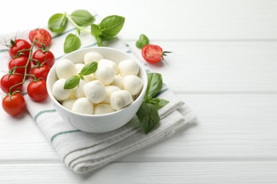 Photo of Tasty mozzarella cheese balls in bowl, basil and tomatoes on white wooden table, space for text