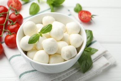 Photo of Tasty mozzarella cheese balls in bowl, basil and tomatoes on white wooden table, closeup