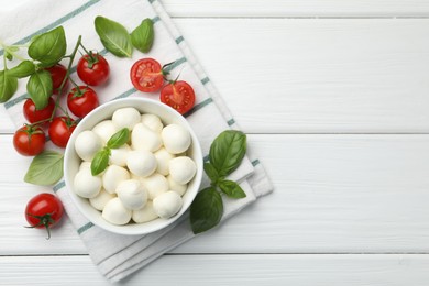Photo of Tasty mozzarella cheese balls in bowl, basil and tomatoes on white wooden table, flat lay. Space for text