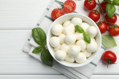 Photo of Tasty mozzarella cheese balls in bowl, basil and tomatoes on white wooden table, flat lay