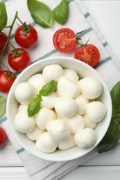 Photo of Tasty mozzarella cheese balls in bowl, basil and tomatoes on white wooden table, flat lay