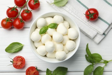 Photo of Tasty mozzarella cheese balls in bowl, basil and tomatoes on white wooden table, flat lay