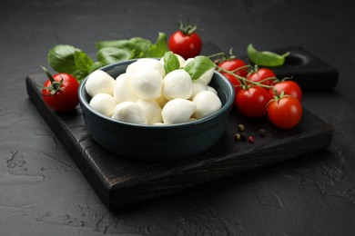 Photo of Tasty mozzarella cheese balls in bowl, basil, peppercorns and tomatoes on black table, closeup