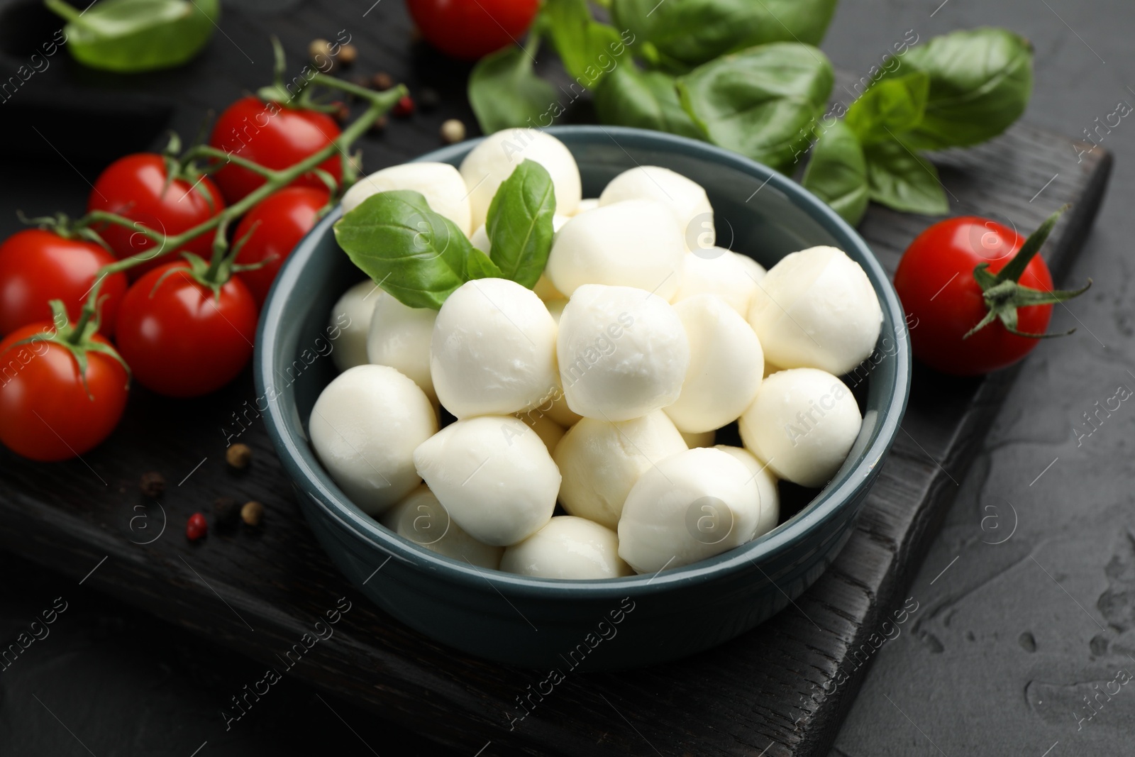Tasty mozzarella cheese balls in bowl, basil, peppercorns and tomatoes on black table, closeup Photo of Tasty mozzarella cheese balls in bowl, basil, peppercorns and tomatoes on black table, closeup