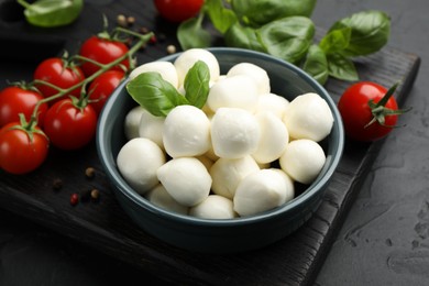 Photo of Tasty mozzarella cheese balls in bowl, basil, peppercorns and tomatoes on black table, closeup