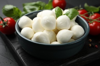 Photo of Tasty mozzarella cheese balls in bowl, basil, peppercorns and tomatoes on black table, closeup