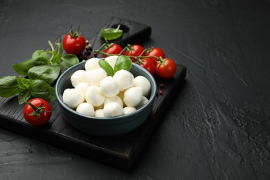 Photo of Tasty mozzarella cheese balls in bowl, basil, peppercorns and tomatoes on black table, closeup. Space for text