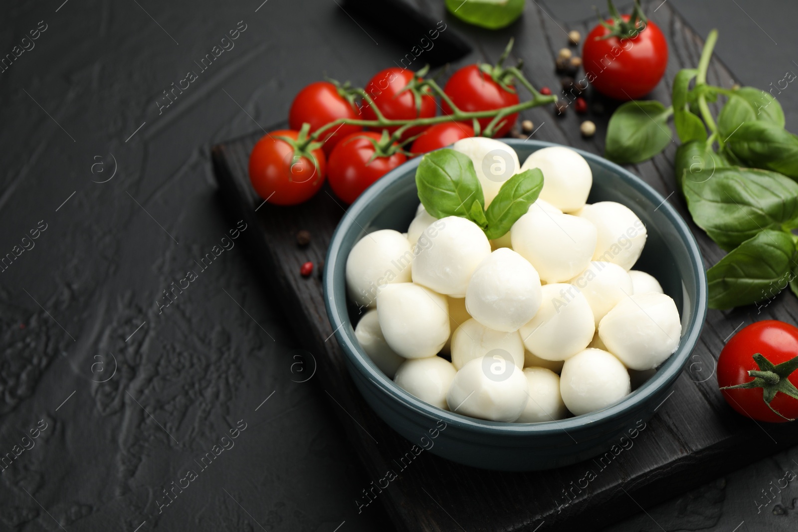 Tasty mozzarella cheese balls in bowl, basil, peppercorns and tomatoes on black table, closeup. Space for text Photo of Tasty mozzarella cheese balls in bowl, basil, peppercorns and tomatoes on black table, closeup. Space for text