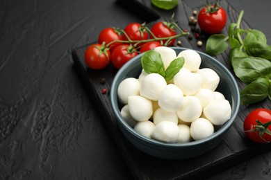Photo of Tasty mozzarella cheese balls in bowl, basil, peppercorns and tomatoes on black table, closeup. Space for text