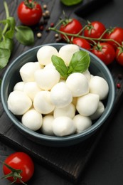 Photo of Tasty mozzarella cheese balls in bowl, basil, peppercorns and tomatoes on black table, above view