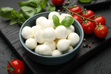 Photo of Tasty mozzarella cheese balls in bowl, basil, peppercorns and tomatoes on black table, closeup