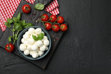 Photo of Tasty mozzarella cheese balls in bowl, basil, peppercorns and tomatoes on black table, top view. Space for text