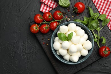 Photo of Tasty mozzarella cheese balls in bowl, basil, peppercorns and tomatoes on black table, top view. Space for text