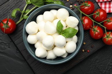 Photo of Tasty mozzarella cheese balls in bowl, basil, peppercorns and tomatoes on black table, top view