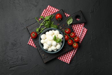 Photo of Tasty mozzarella cheese balls in bowl, basil, peppercorns and tomatoes on black table, top view