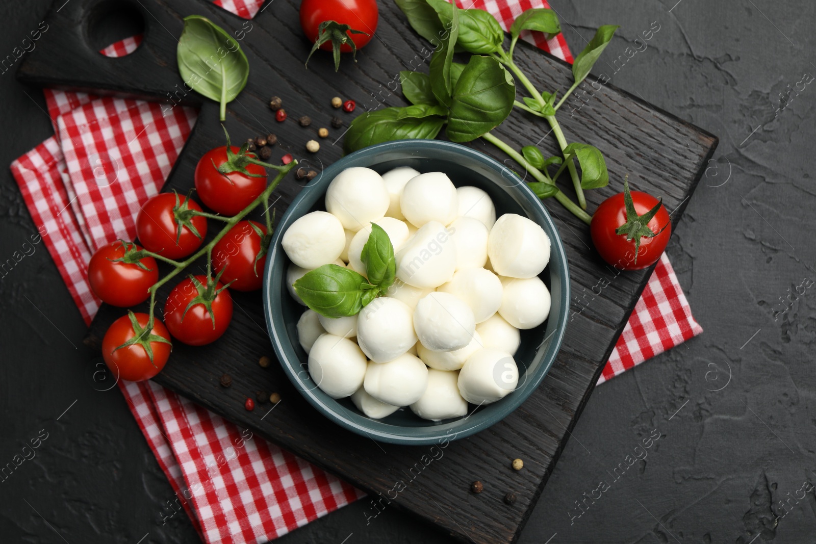 Tasty mozzarella cheese balls in bowl, basil, peppercorns and tomatoes on black table, top view Photo of Tasty mozzarella cheese balls in bowl, basil, peppercorns and tomatoes on black table, top view