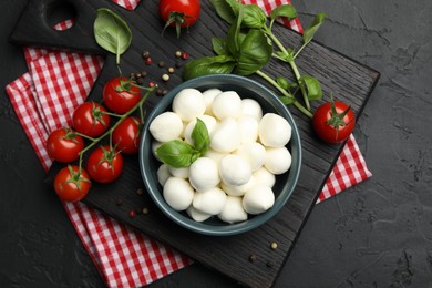 Photo of Tasty mozzarella cheese balls in bowl, basil, peppercorns and tomatoes on black table, top view