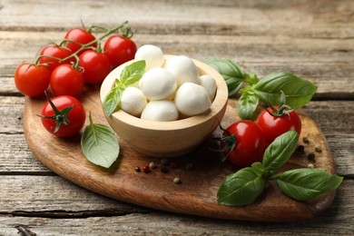 Photo of Tasty mozzarella cheese balls in bowl, basil, peppercorns and tomatoes on wooden table, closeup