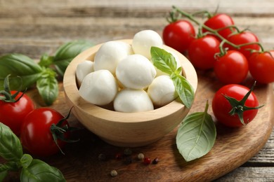 Photo of Tasty mozzarella cheese balls in bowl, basil, peppercorns and tomatoes on wooden table, closeup