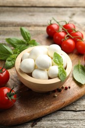 Photo of Tasty mozzarella cheese balls in bowl, basil, peppercorns and tomatoes on wooden table, closeup