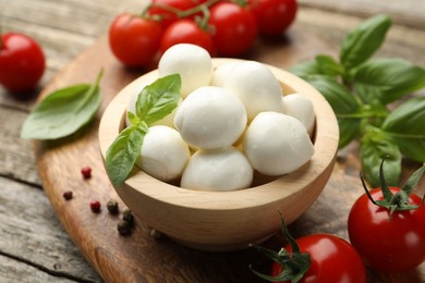 Photo of Tasty mozzarella cheese balls in bowl, basil, peppercorns and tomatoes on wooden table, closeup