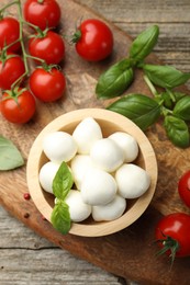 Photo of Tasty mozzarella cheese balls in bowl, basil, peppercorns and tomatoes on wooden table, top view