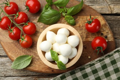 Photo of Tasty mozzarella cheese balls in bowl, basil, peppercorns and tomatoes on wooden table, top view
