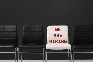 Image of White chair with phrase We Are Hiring and empty black ones near dark wall. Recruitment, human resource concept