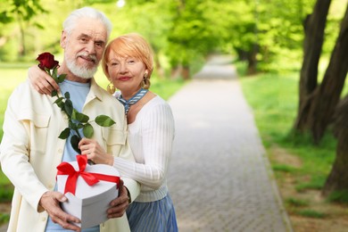 Photo of Lovely senior couple with gift box and rose in park, space for text