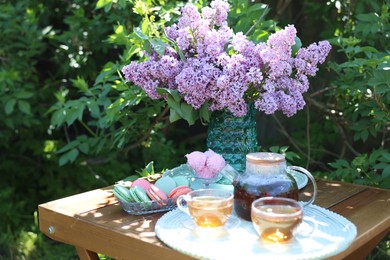 Photo of Beautiful lilac flowers in vase, sweets and tea on wooden table in garden