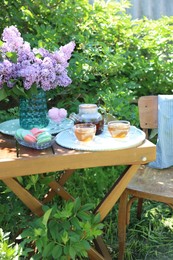 Photo of Beautiful lilac flowers in vase, sweets and tea on wooden table in garden