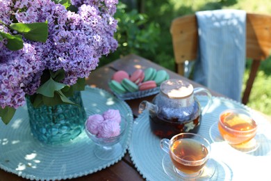 Photo of Beautiful lilac flowers in vase, sweets and tea on table in garden, closeup