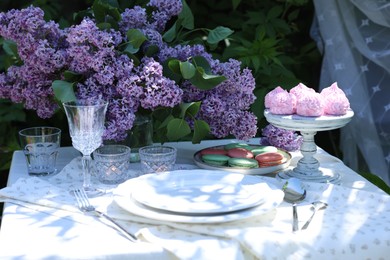 Photo of Beautiful lilac flowers, sweets, plates, cutlery, glasses and burning candles on table in garden