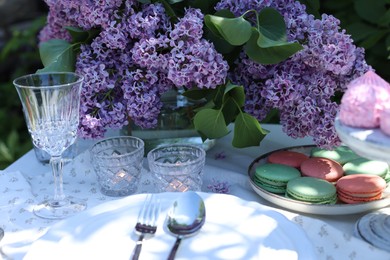 Photo of Beautiful lilac flowers, sweets, plates, cutlery, glass and burning candles on table in garden, closeup