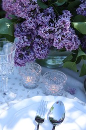 Photo of Beautiful lilac flowers, plates, cutlery, glass and burning candles on table in garden, closeup
