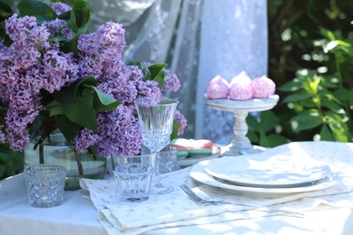 Photo of Beautiful lilac flowers, sweets, plates, cutlery and glasses on table in garden
