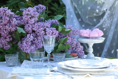 Photo of Beautiful lilac flowers, sweets, plates, cutlery and glasses on table in garden